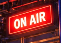 THE ON AIR SIGN. The illuminated sign marks the threshold of live television. It anchors the scene to the broadcast moment, while the exact sign and backstage view are illustrative inventions.