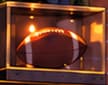THE FOOTBALL CASE. A football rests in a clear display case beneath the television. Together with the framed sports pictures, it establishes the fictional bar's atmosphere. It is an invented prop, with no claimed signature or match provenance.