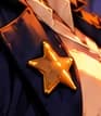 THE SHERIFF'S STAR. A plain gold star catches the sunset against the navy coat. The new sheriff gives regulatory oversight a familiar frontier face.